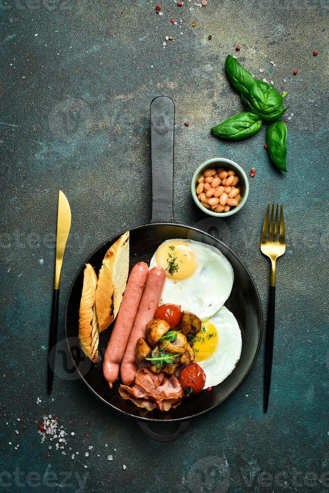 Traditional American breakfast fried eggs, sausage, bacon and vegetables in a frying pan. Homemade food. On a dark stone background. photo