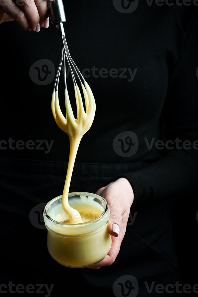 Fresh homemade sweetened condensed milk in a jar and a kitchen whisk. On a black background. photo