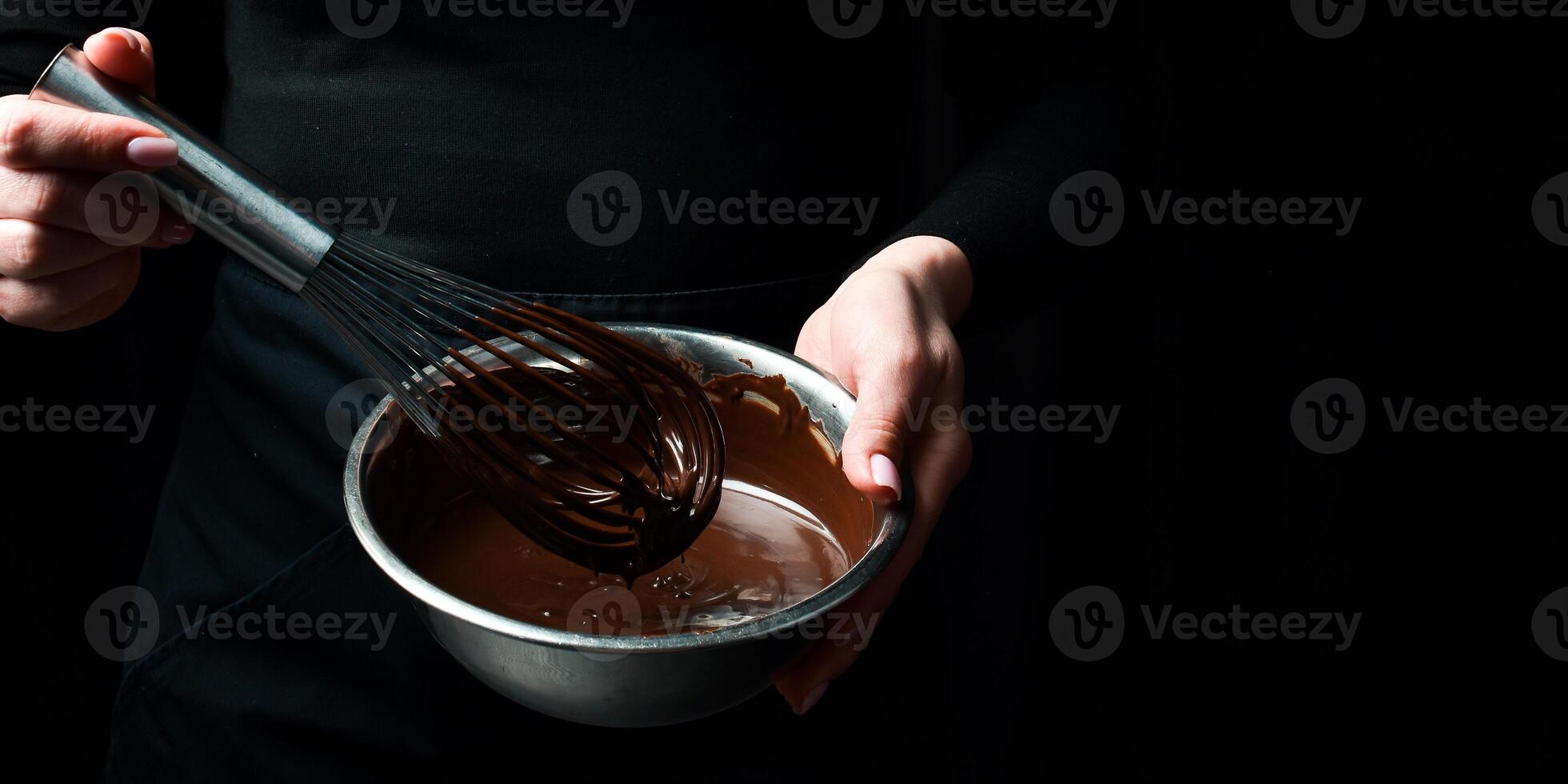 A bowl of melted chocolate and a kitchen whisk are held by hands. Kitchen utensils. On a black background. photo