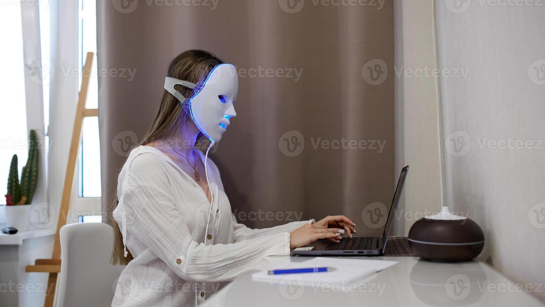A beautiful girl with an LED mask on her head works at a laptop. Home skin care concept. photo