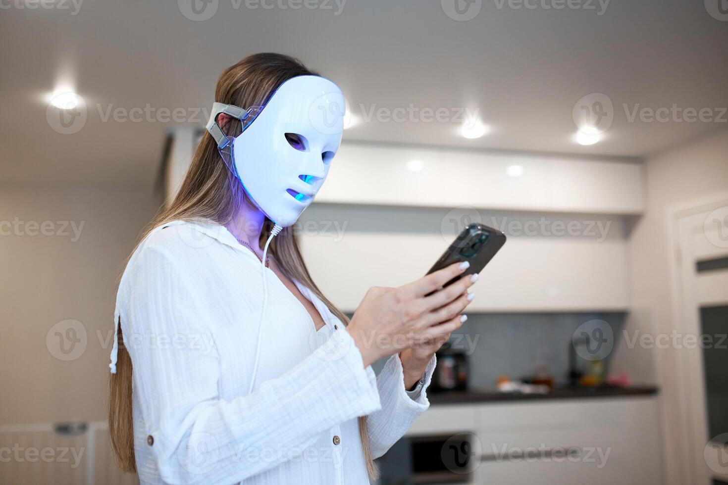 Portrait of a beautiful woman communicating on the phone and performing a light therapy facial mask procedure at home. photo