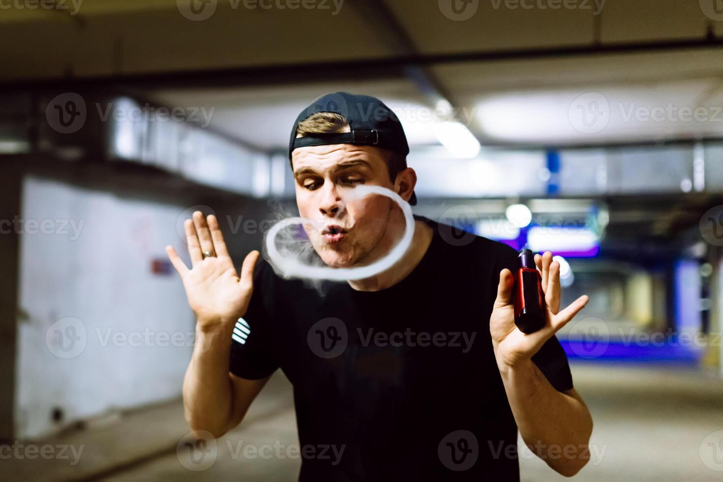 Man in cap smoke an electronic cigarette and releases clouds of vapor performing various kind of vaping tricks photo