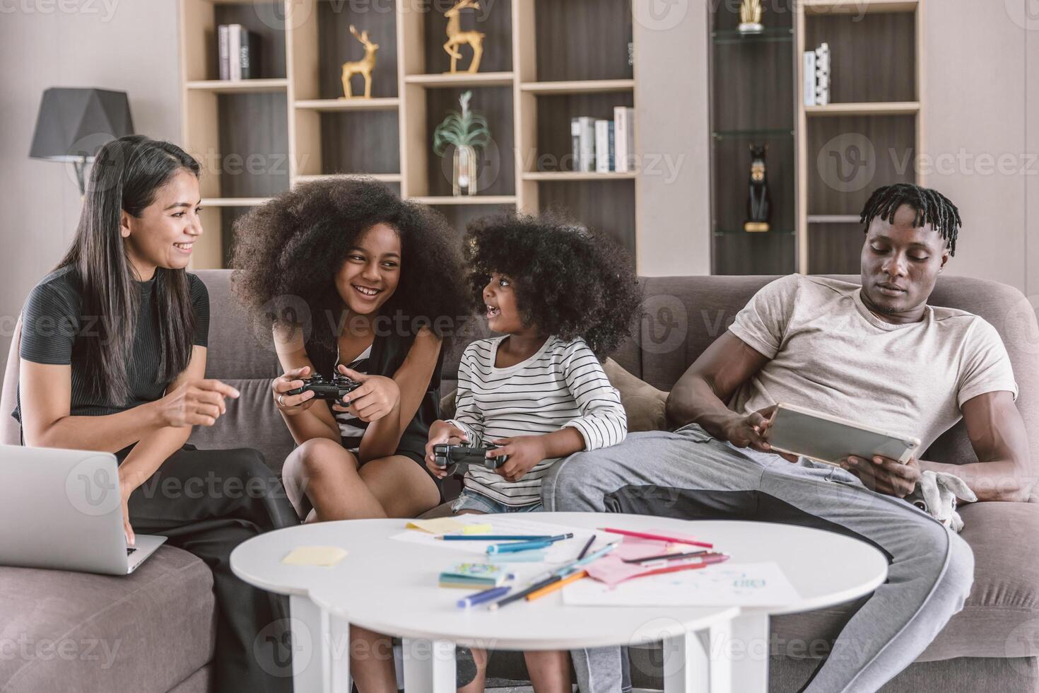 Happy black african family enjoy holiday together at sofa living room two daughter playing game mother online working with laptop computer and dad looking at tablet photo