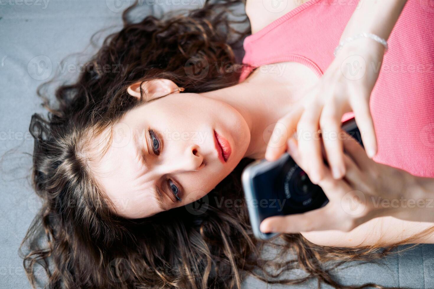 joven mujer con largo Rizado pelo mentiras en el sofá y navega social redes en su teléfono con un grave mirar, parte superior ver foto