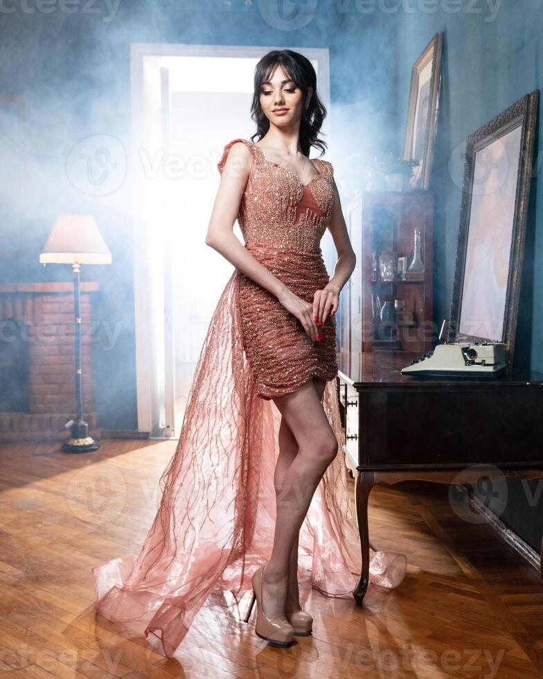 A woman in a pink dress stands in front of a vintage desk and looks into the camera. photo