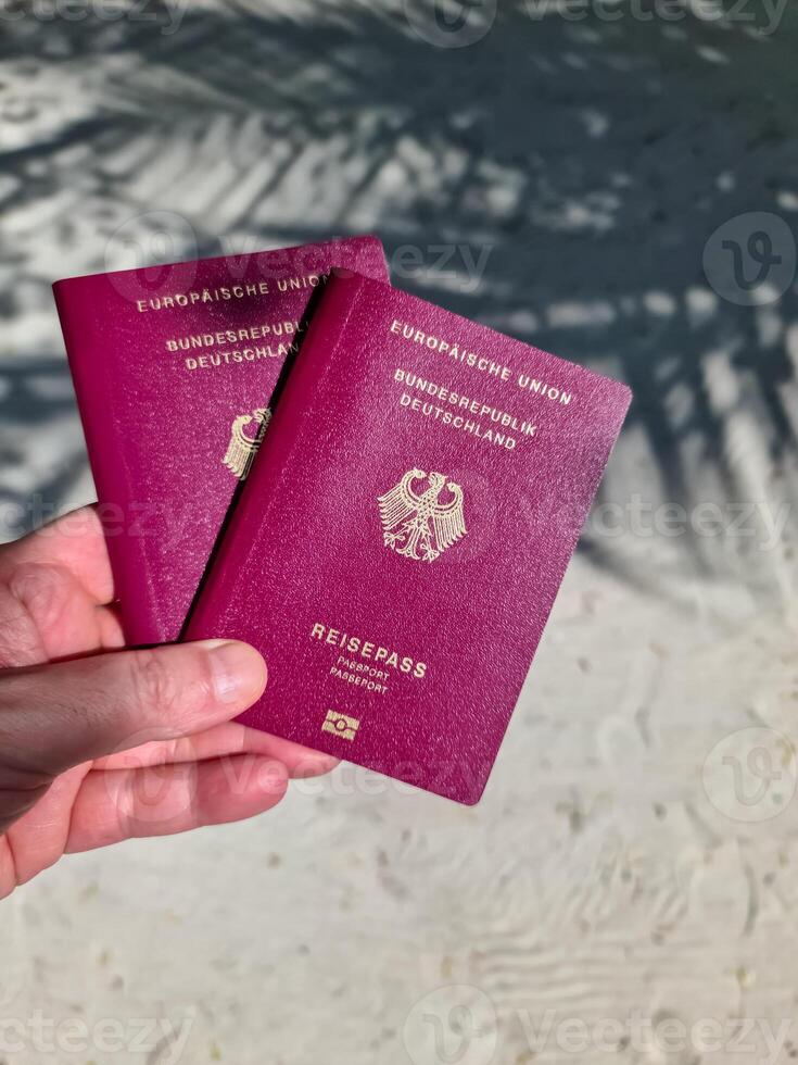 A hand holds two German passports in front of a soft travel background in the Maldives with palm trees and beach. photo