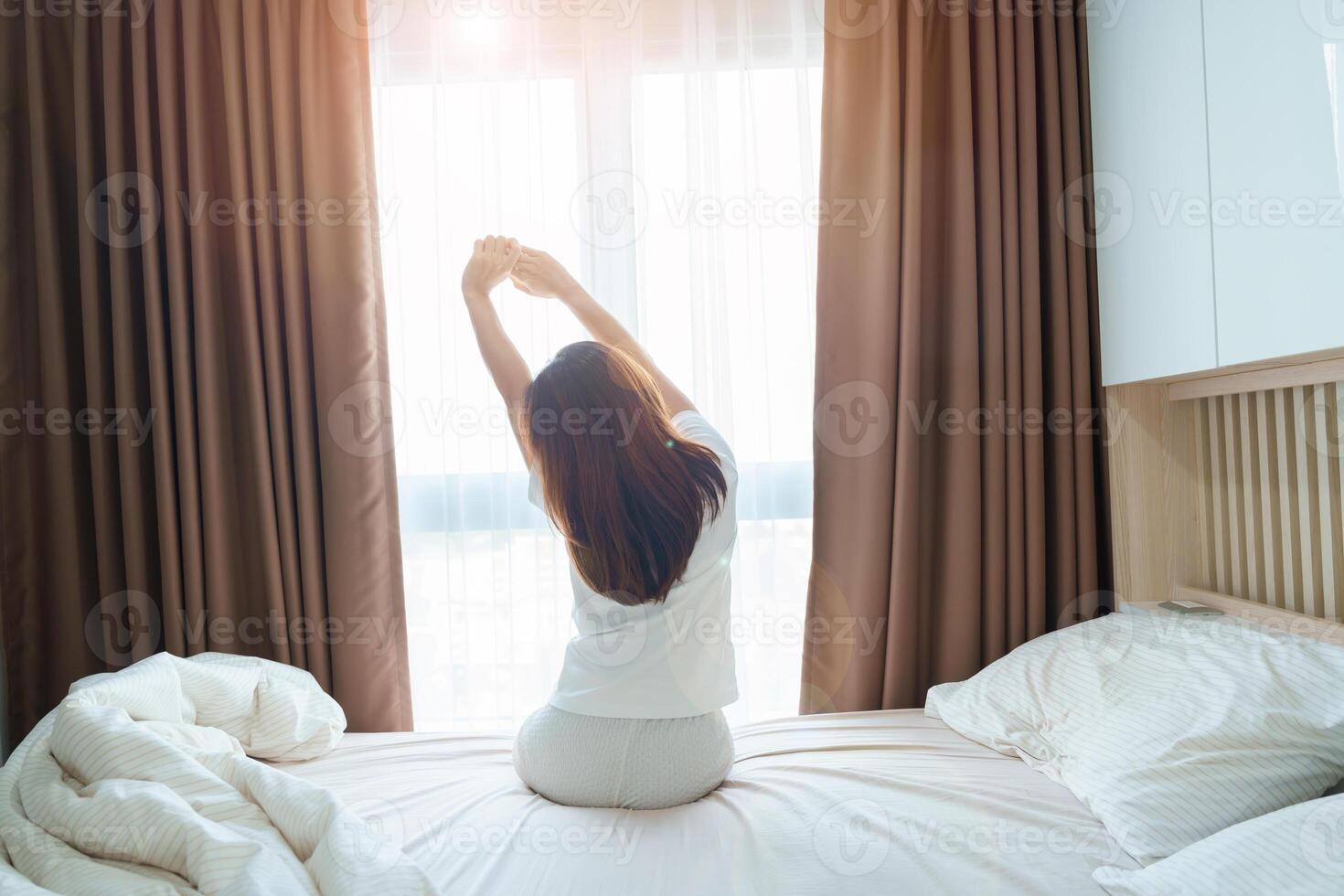 happy woman stretching on bed after wake up, young adult female rising arms and looking to window in the early morning. fresh relax and have a nice day concepts photo