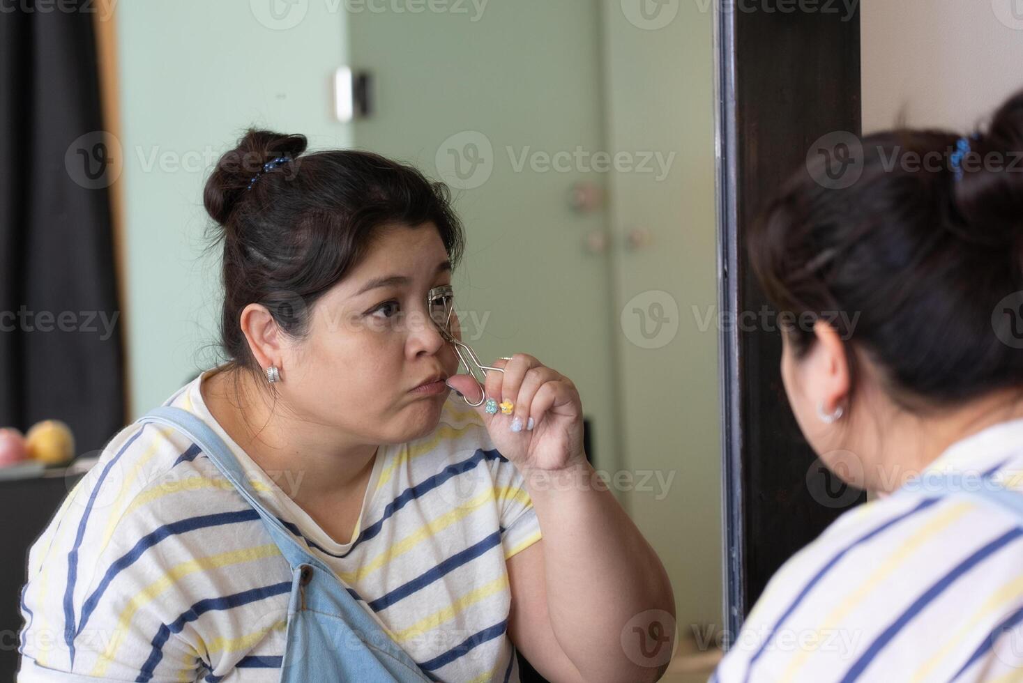 Reflection of Beautiful Asian Plus-size Woman Using Large Mirror to Curl Eyelashes with Curler photo