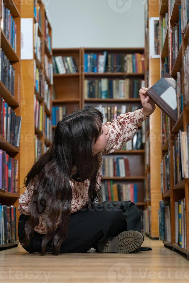 Student girl, sitting on the floor in the library and choosing book for knowledge, learning or literature at book store. Female student sitting on floor by bookshelf in study, research or education photo
