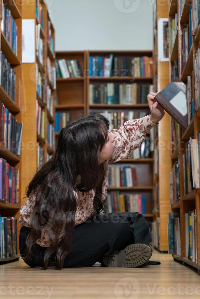 estudiante chica, sentado en el piso en el biblioteca y elegir libro para conocimiento, aprendizaje o literatura a libro almacenar. hembra estudiante sentado en piso por estante para libros en estudiar, investigación o educación foto