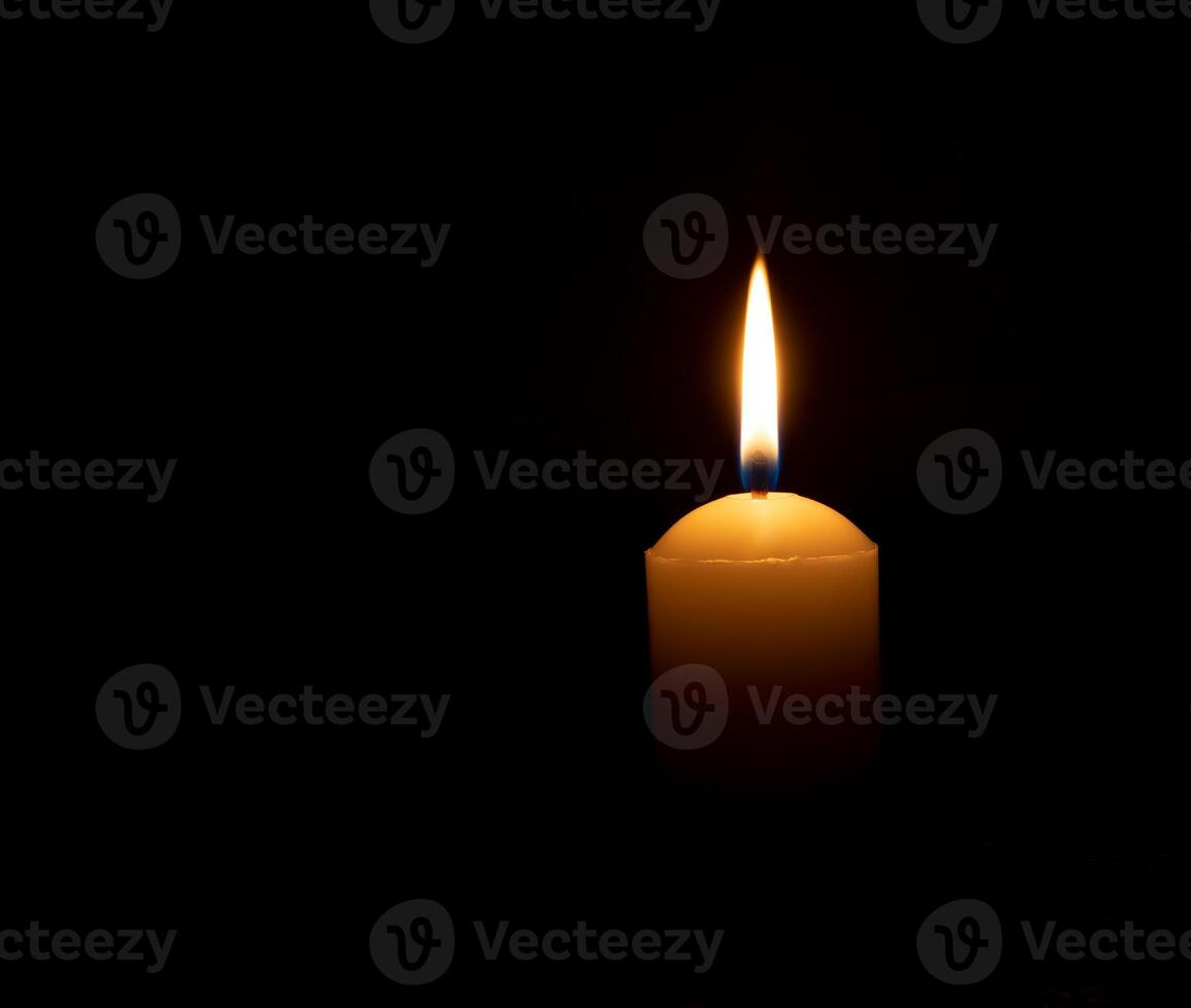 Single burning candle flame or light glowing on a big white candle on black or dark background on table in church for Christmas, funeral or memorial service with copy space. photo