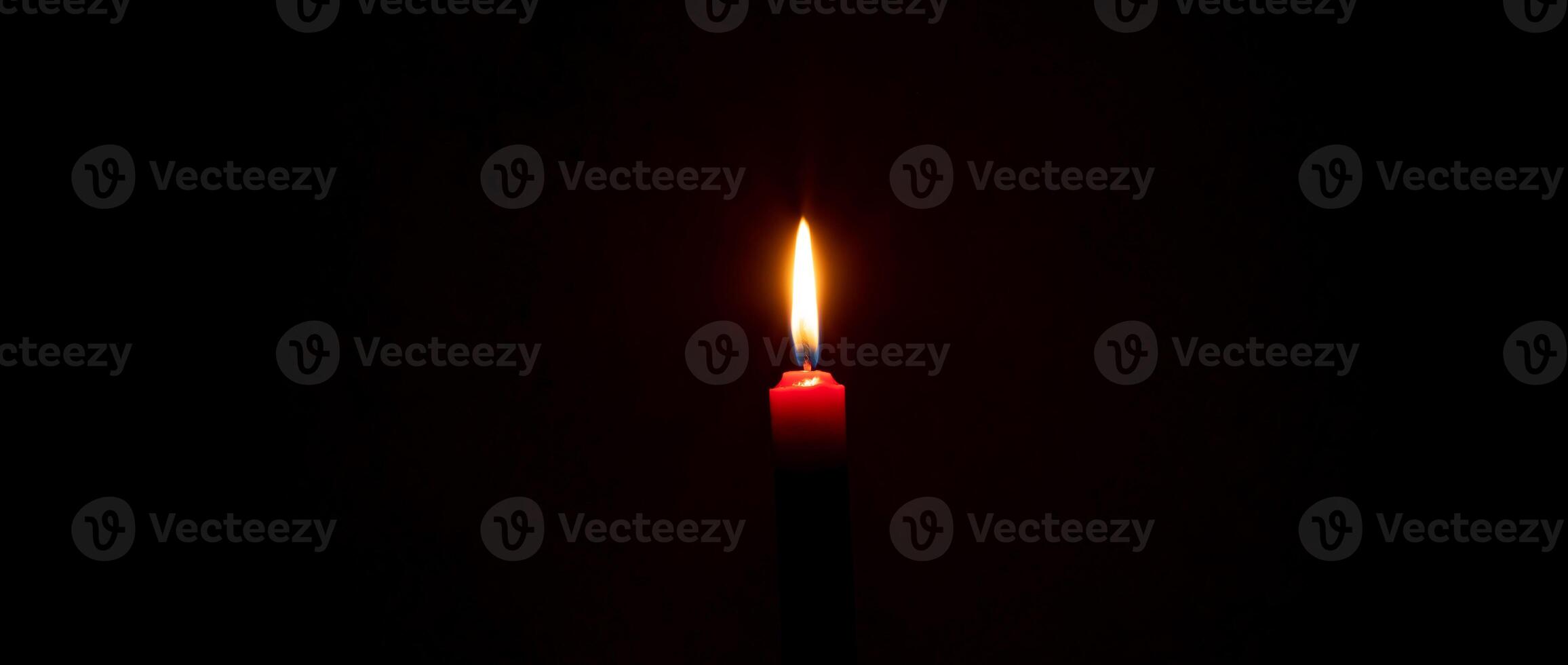 Single burning candle flame or light glowing on a small red candle on black or dark background on table in church for Christmas, funeral or memorial service with copy space photo