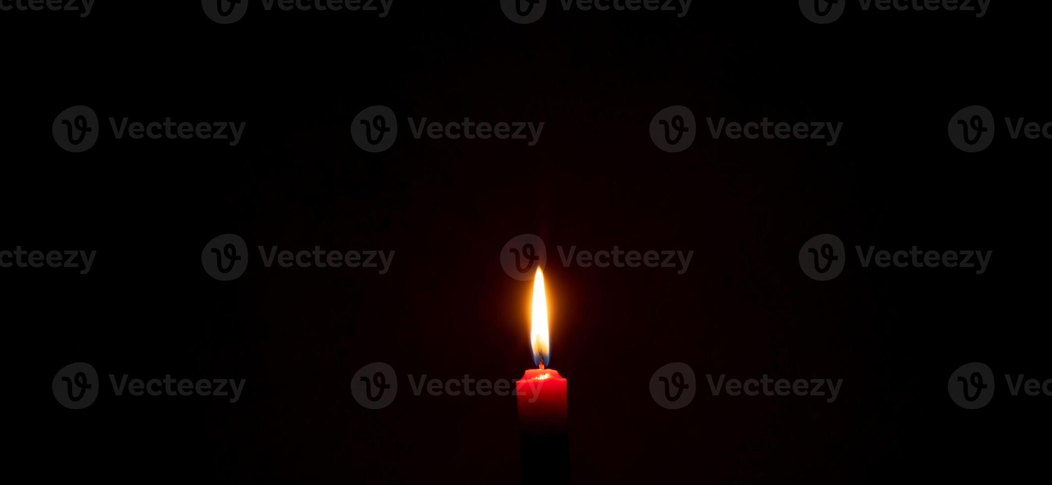 A single burning candle flame or light glowing on a red candle on black or dark background on table in church for Christmas, funeral or memorial service with copy space photo