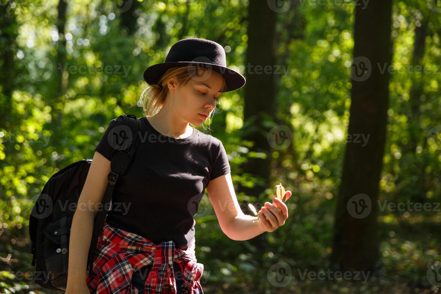 young caucasian woman in hat and with backpack is in the forest and looks at the compass in her hand.background blurred. selective focus. High quality photo