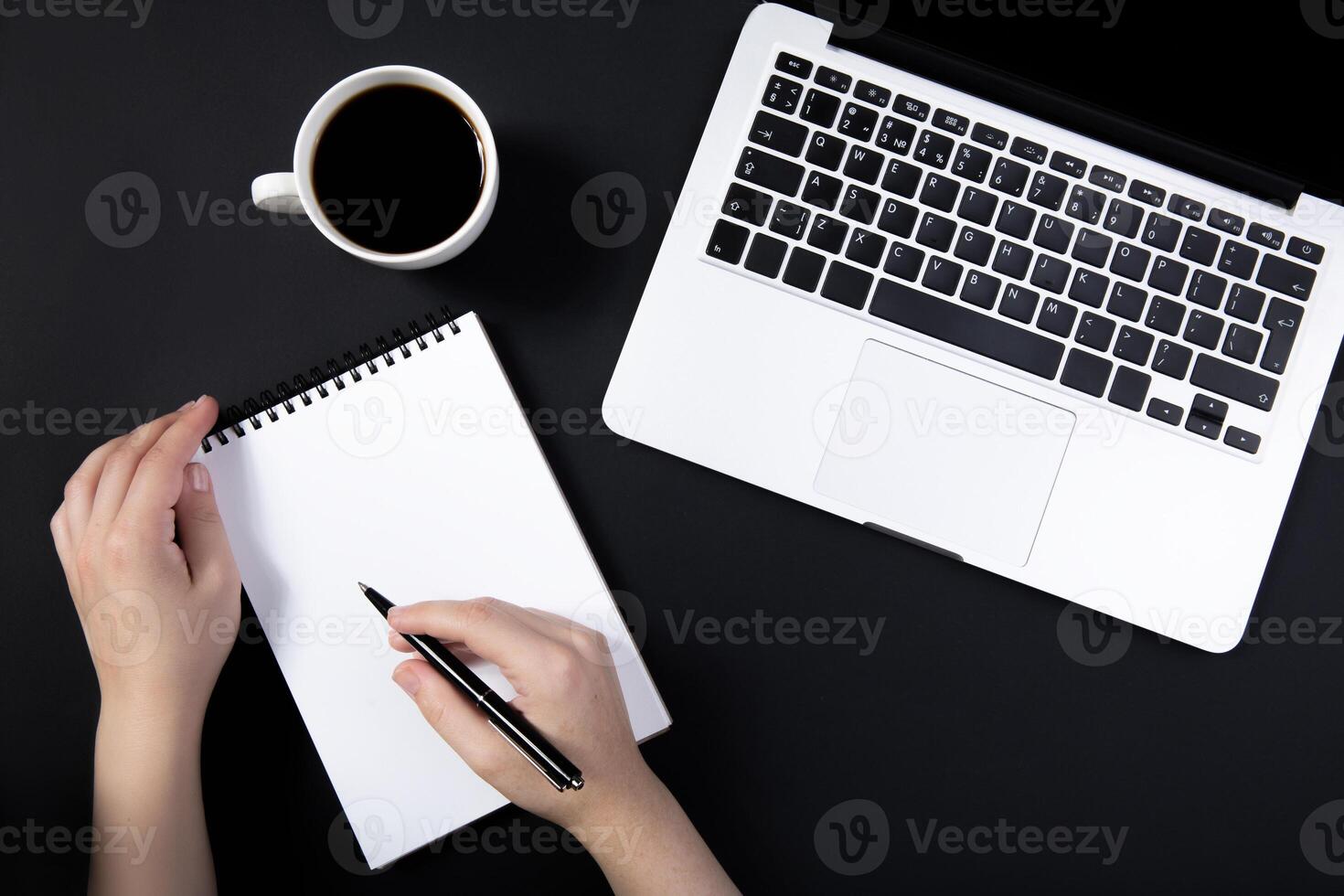 hand holds pen over notebook on black table near laptop. cup of coffee nearby. Top view with copy space for text input. photo