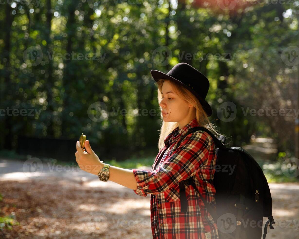young caucasian woman in hat and with backpack is in the forest and looks at the compass in her hand.background blurred. selective focus. High quality photo