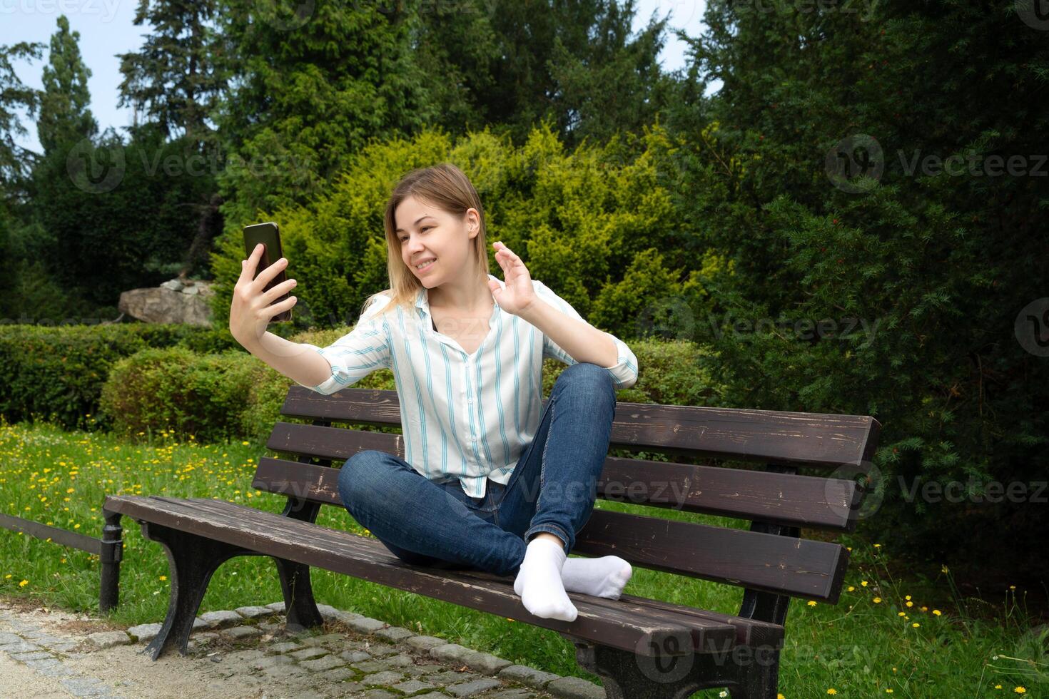 happy Caucasian woman sitting on a park bench on a sunny day, holding a phone in her hand, talking on video chat and waving her hand photo