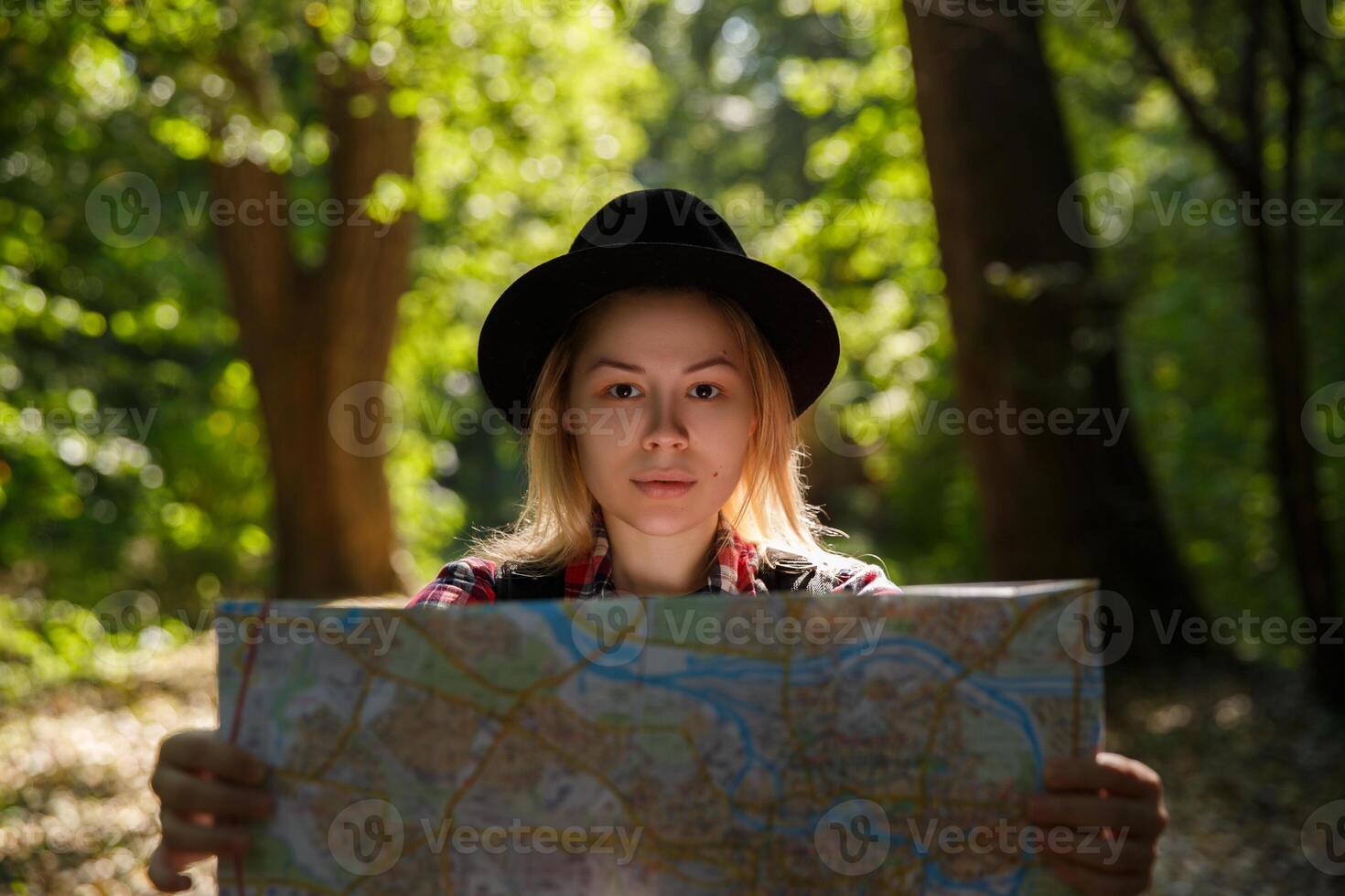 young caucasian woman in a hat looking at a geographic map in the shade of trees in a park or forest. High quality photo