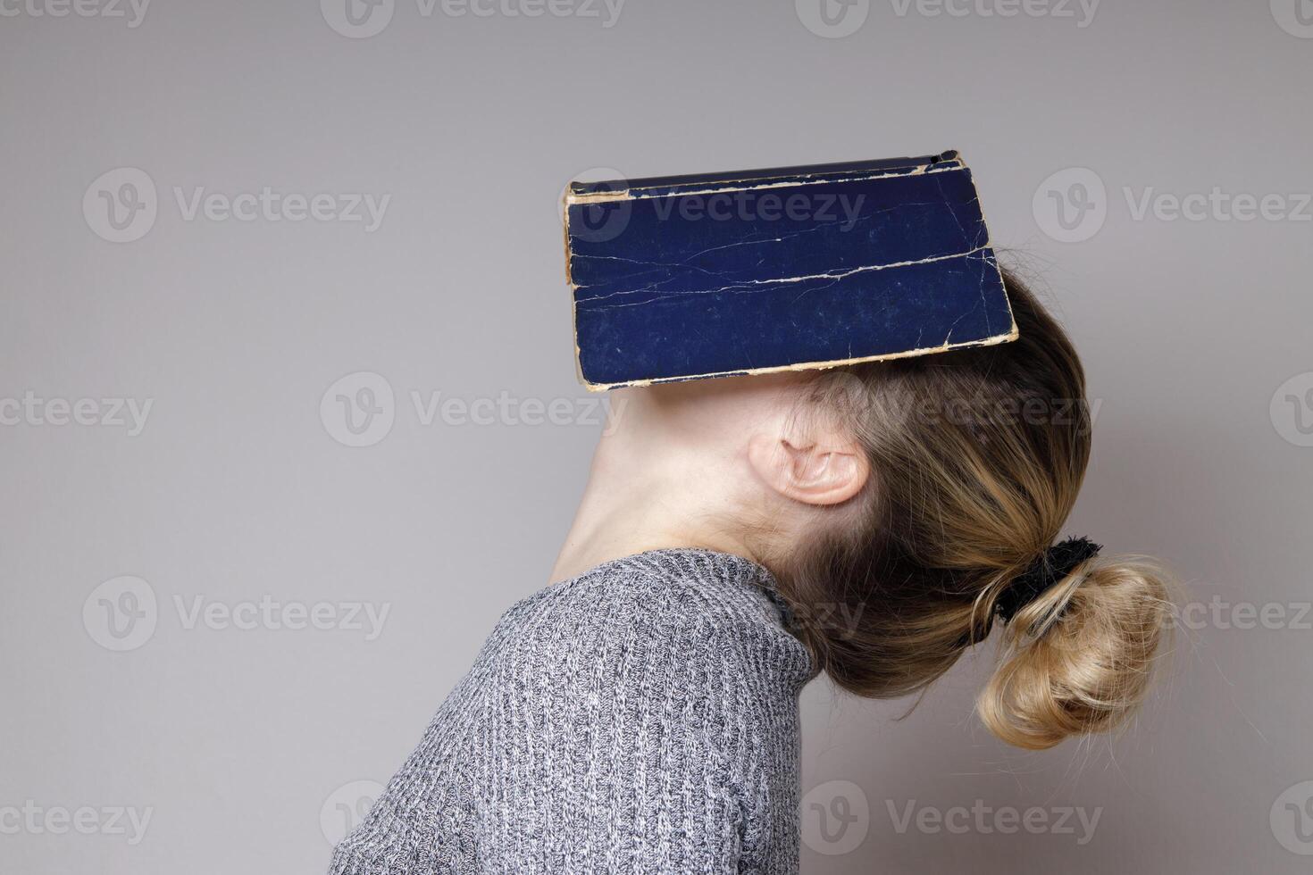 Horizontal image of a young woman covering her face with a book. A beautiful woman in a gray knitted sweater, hiding her face behind a blue book cover, with her head thrown back. Book concept photo