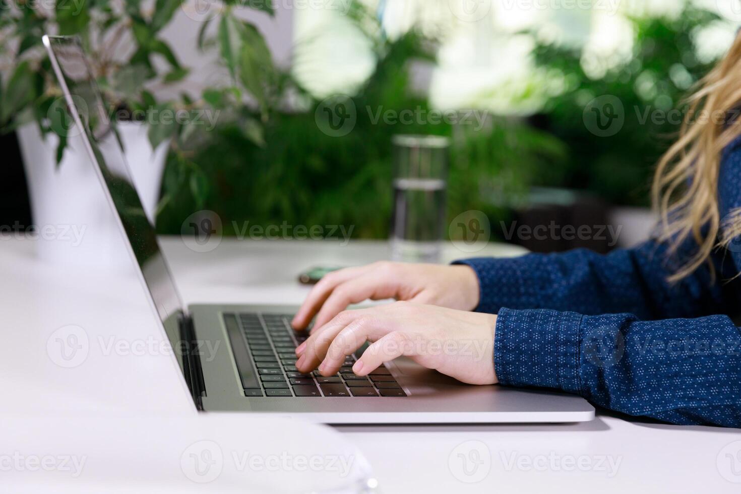 el manos de un invisible niña en un azul blusa son mecanografía en un ordenador portátil a el mesa. allí es un móvil teléfono cerca y allí son muchos flores en un maceta. alto calidad foto