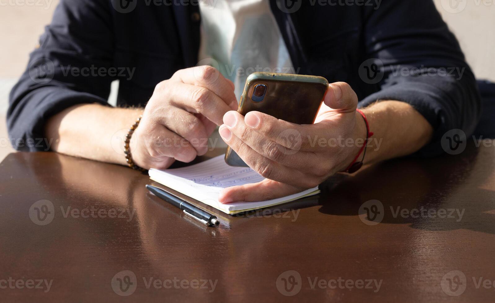 an unknown man holds a phone in one hand, he touches the screen with the finger of the other hand, a notepad and a pen lie on a brown wooden table nearby. High quality photo