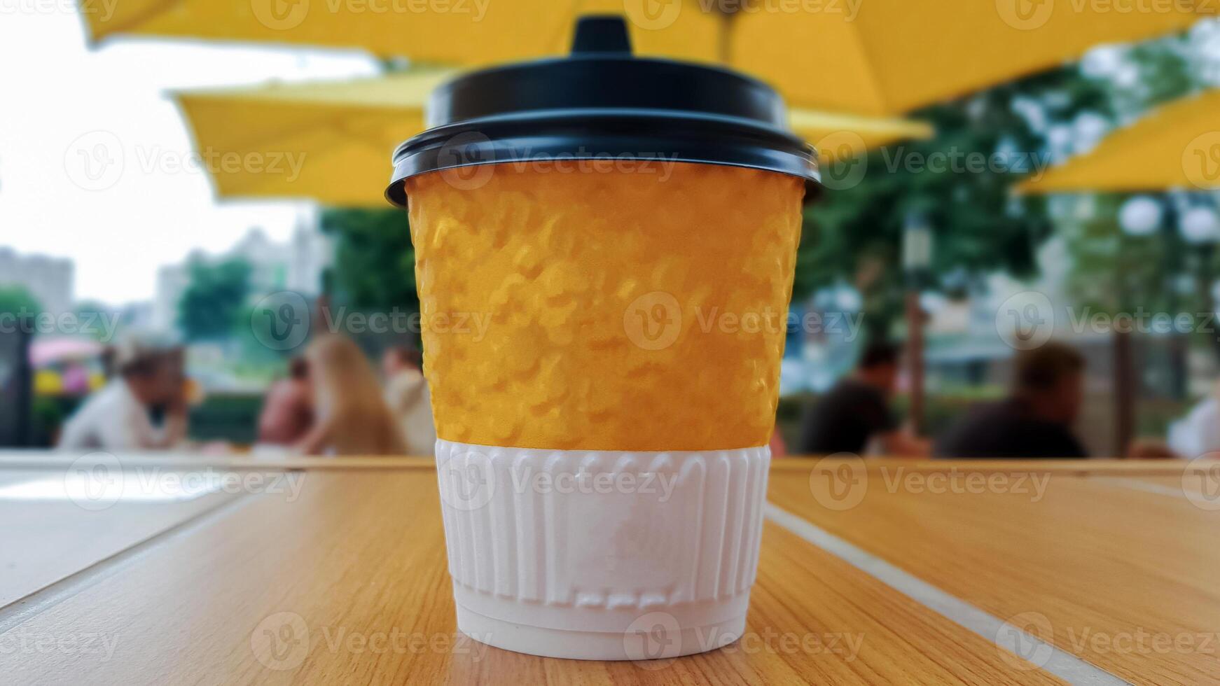 One yellow paper glass of tea or coffee - takeaway food on a table in a coffee shop. Nobody. A disposable glass stands against the backdrop of a summer cafe with unrecognizable people. photo
