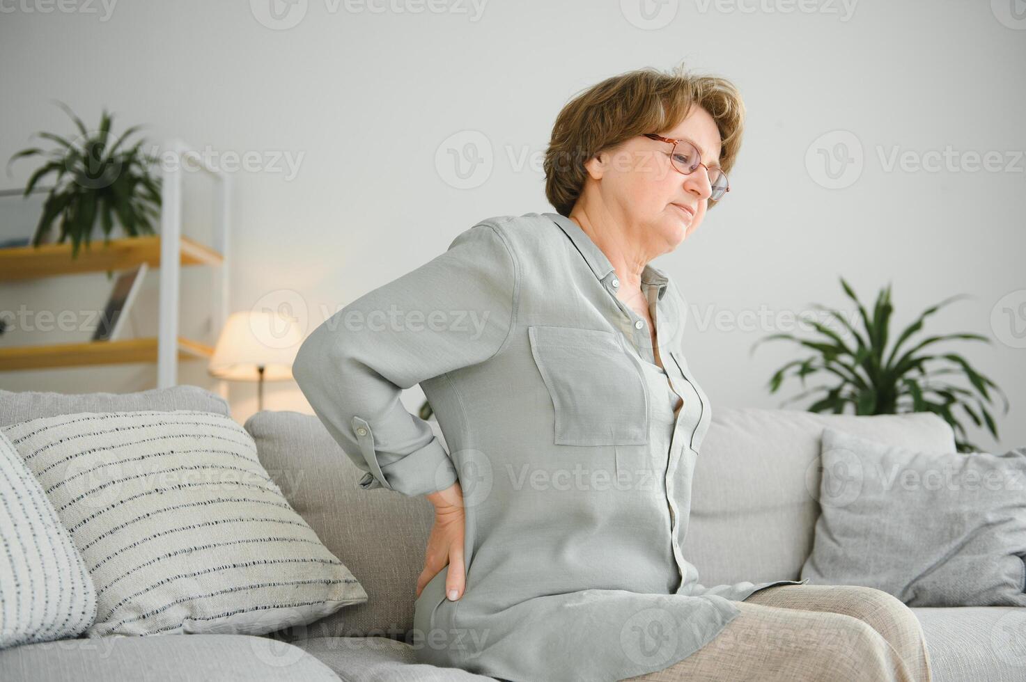maduro dama sentado en cama y conmovedor su atrás, radiculitis y dolor foto