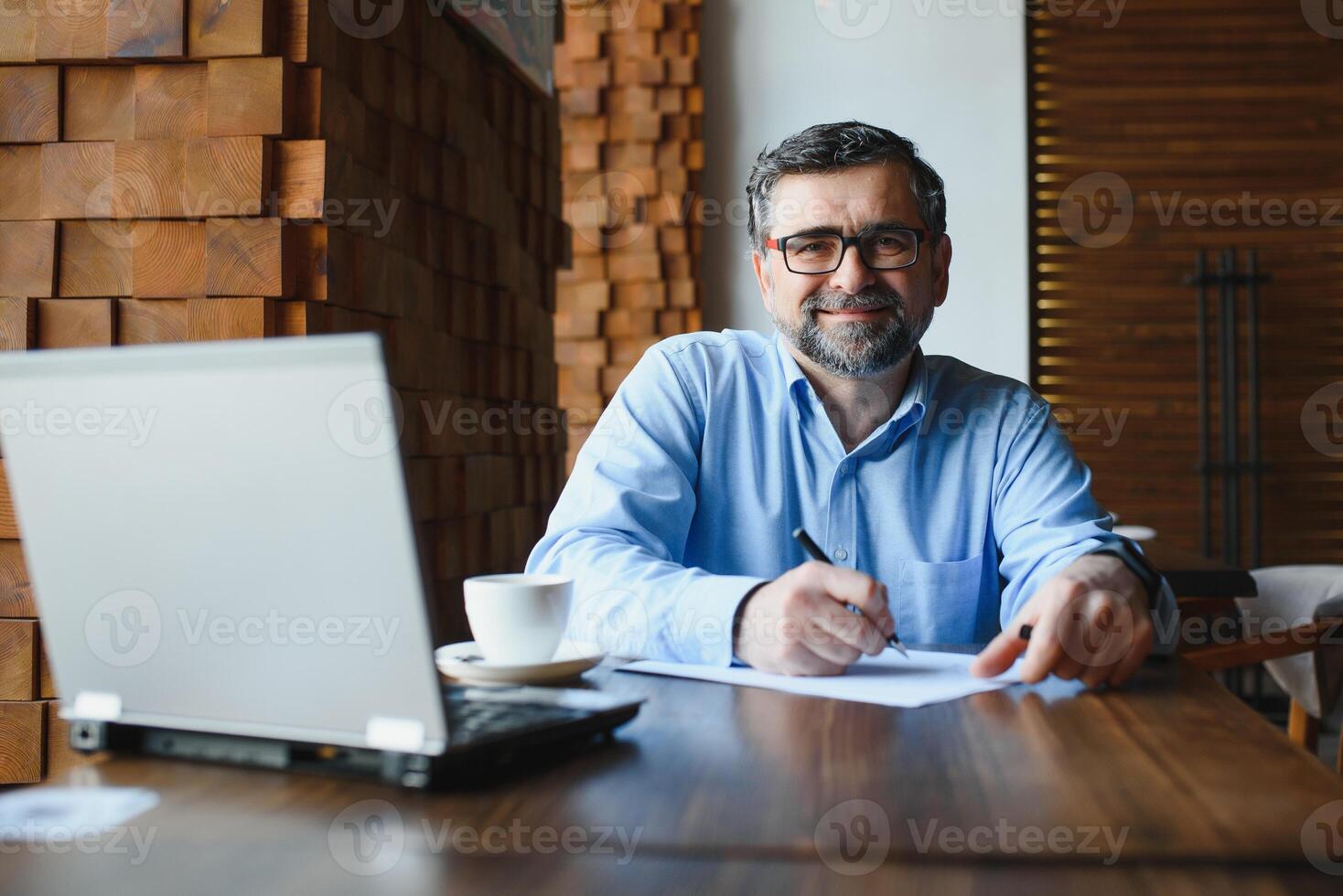 Mature businessman drinking coffee in cafe. Portrait of handsome man wearing stylish eyeglasses using laptop, looking at camera, smiling. Coffee break concept photo