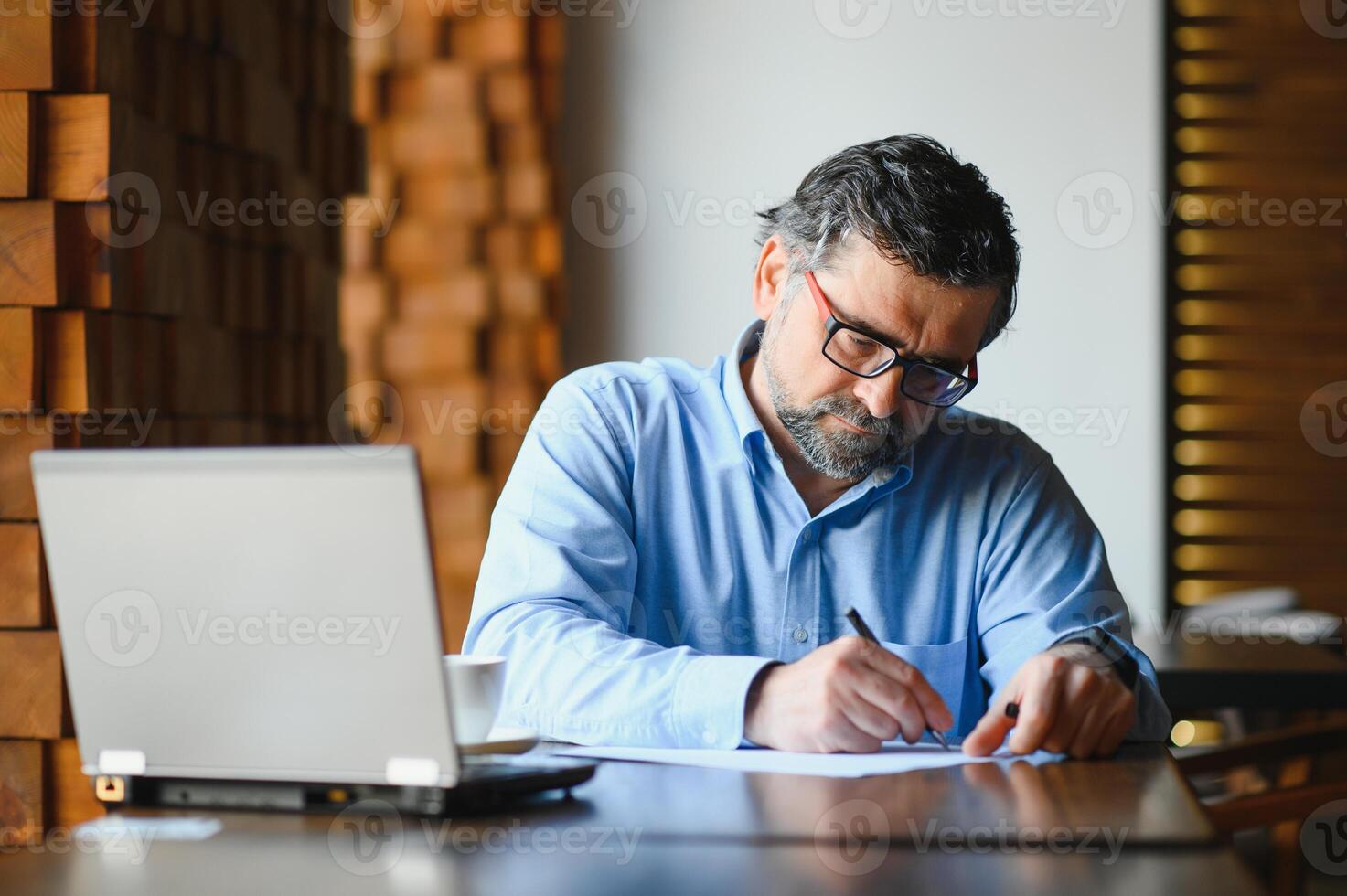 Positive senior bearded man with grey hair drinking coffee and using laptop at cafe, copy space. Stylish aged businessman in burgundy jacket enjoying his tea while working online. photo