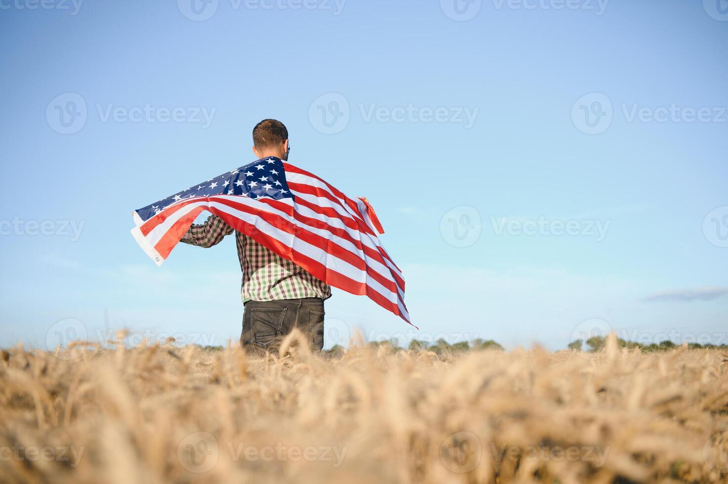 Young patriotic farmer stands among new harvest. Boy walking with the american flag on the wheat field celebrating national independence day. 4th of July concept. photo