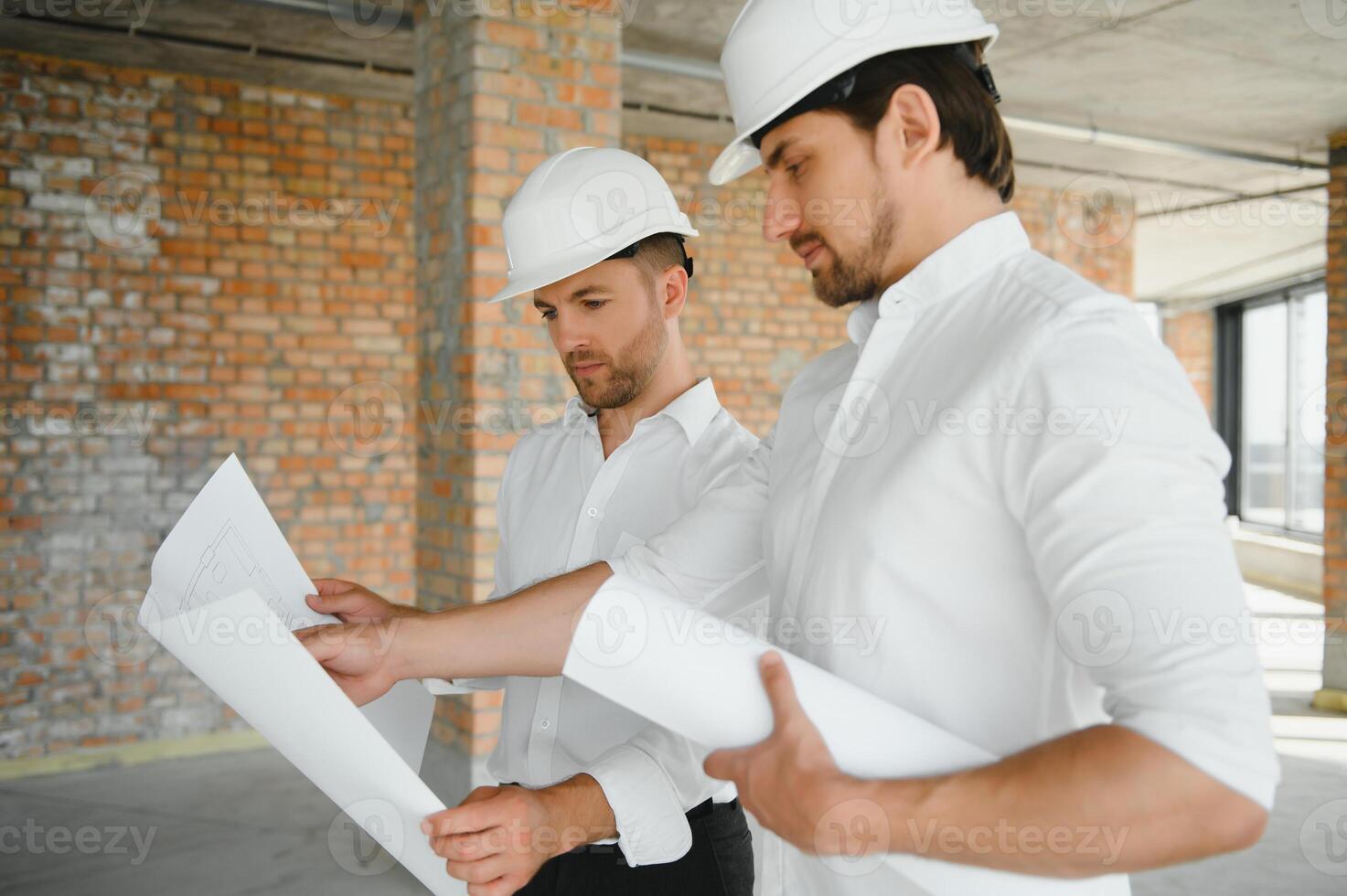 A front view of two smart architects with white helmets reviewing blueprints at a construction site on a bright sunny day photo