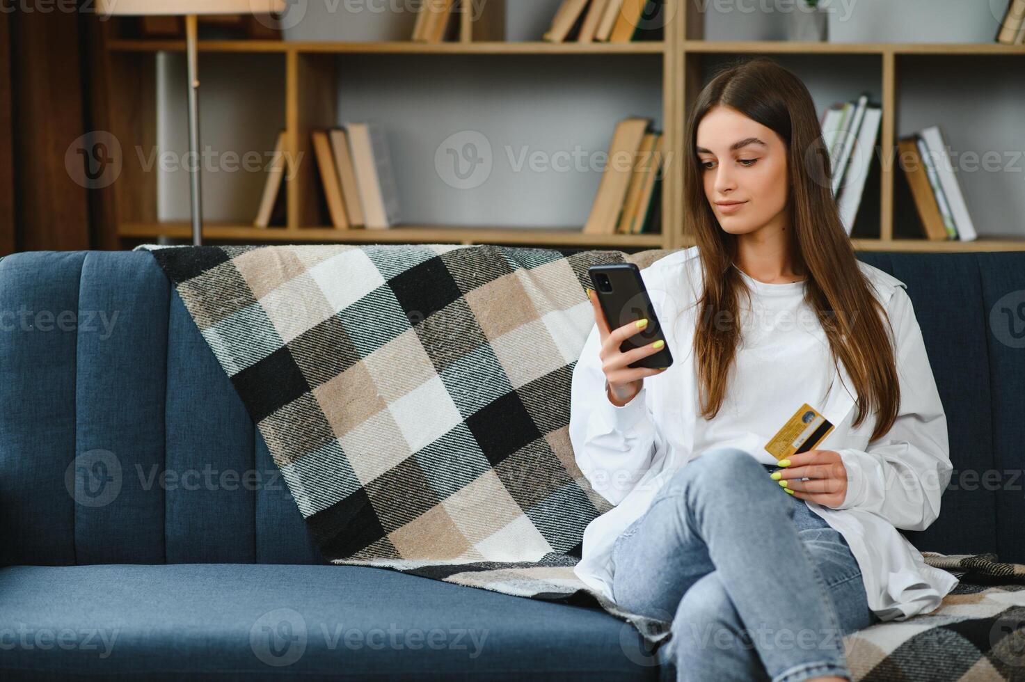 Happy young woman holding phone and credit card sitting on the couch. Online shopping. photo