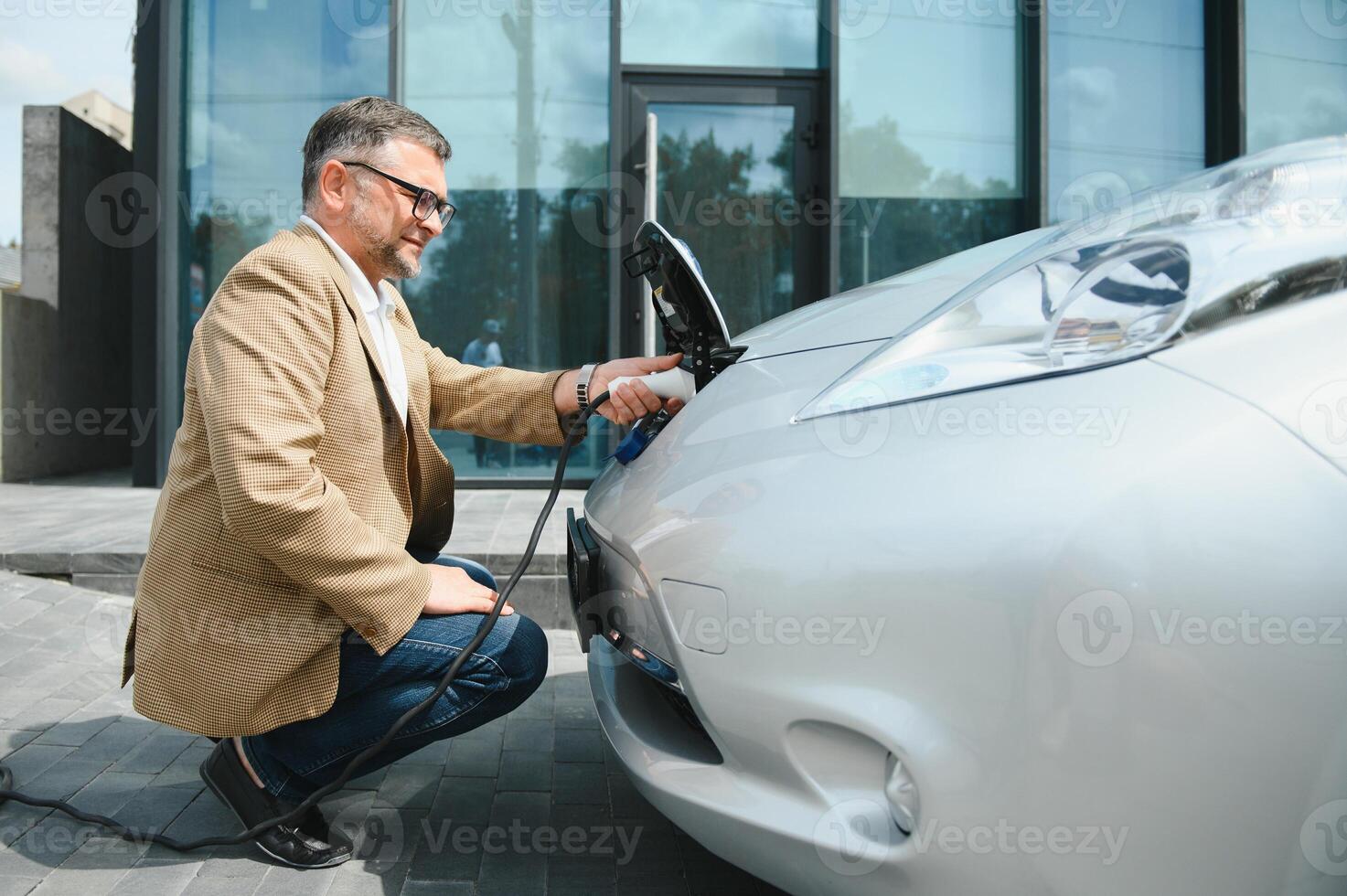 hansome barbado hombre sentado cerca su nuevo moderno eléctrico coche y participación enchufe de el cargador, mientras coche es cargando a el cargando estación foto