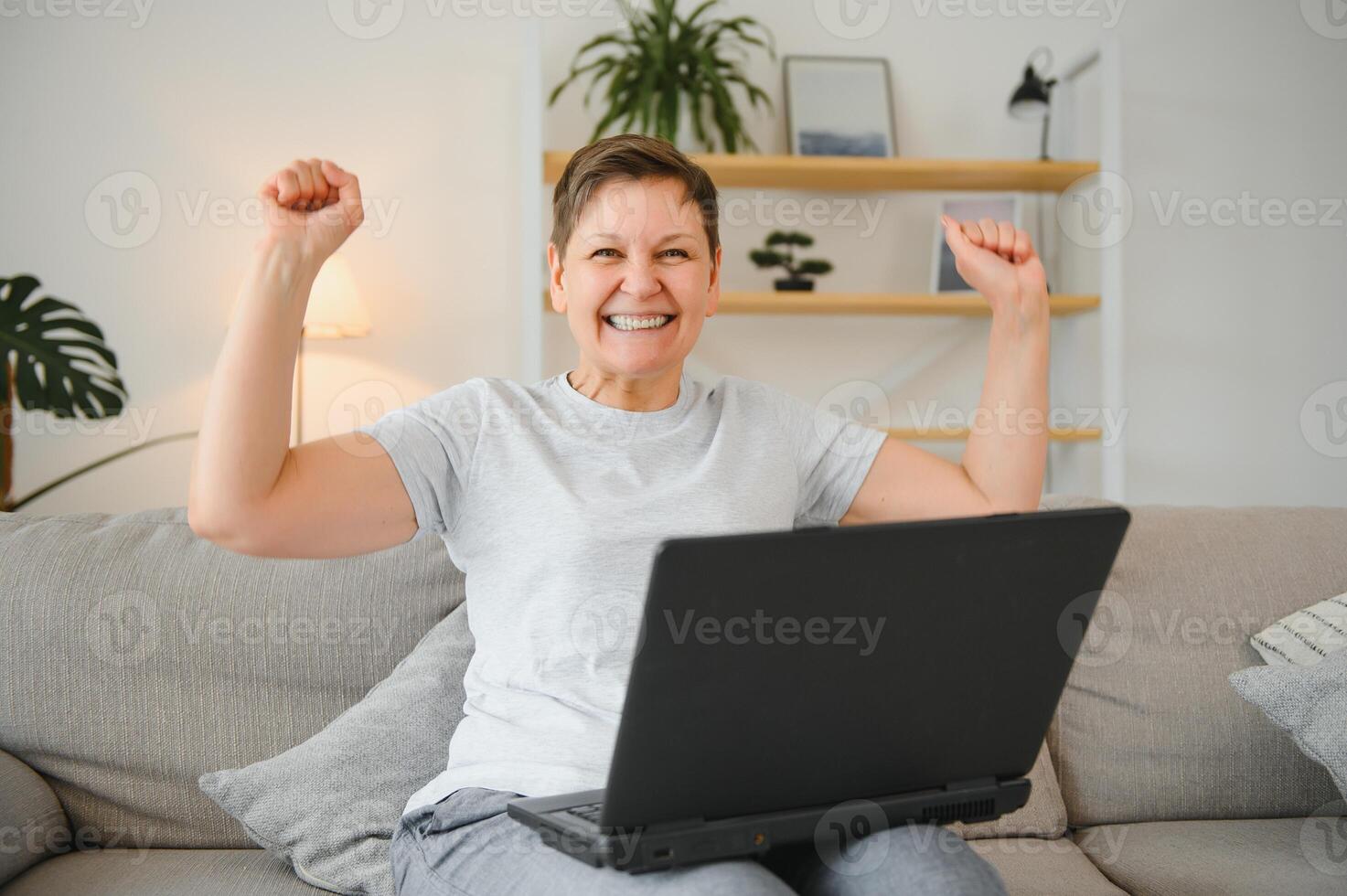 Excited mature woman looking at laptop screen, reading good news in message, celebrating online lottery win, rejoicing success, overjoyed older female sitting on couch at home, using computer. photo