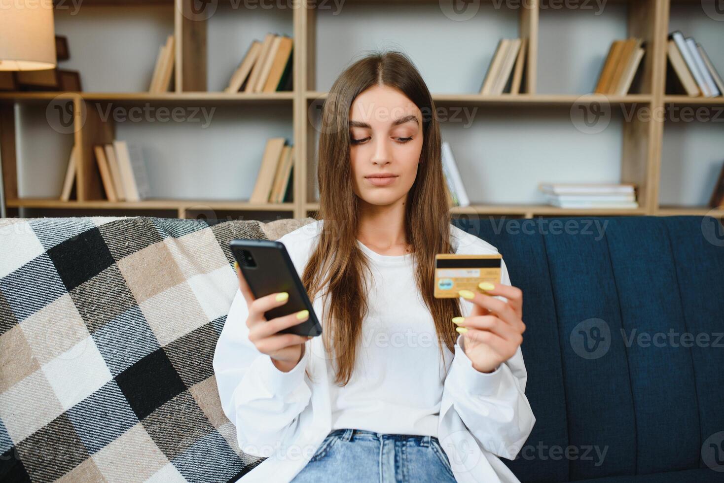 contento adolescente haciendo compras en línea sentado en sofá a hogar. joven mujer insertar crédito tarjeta número en sitio web con su inteligente teléfono. foto