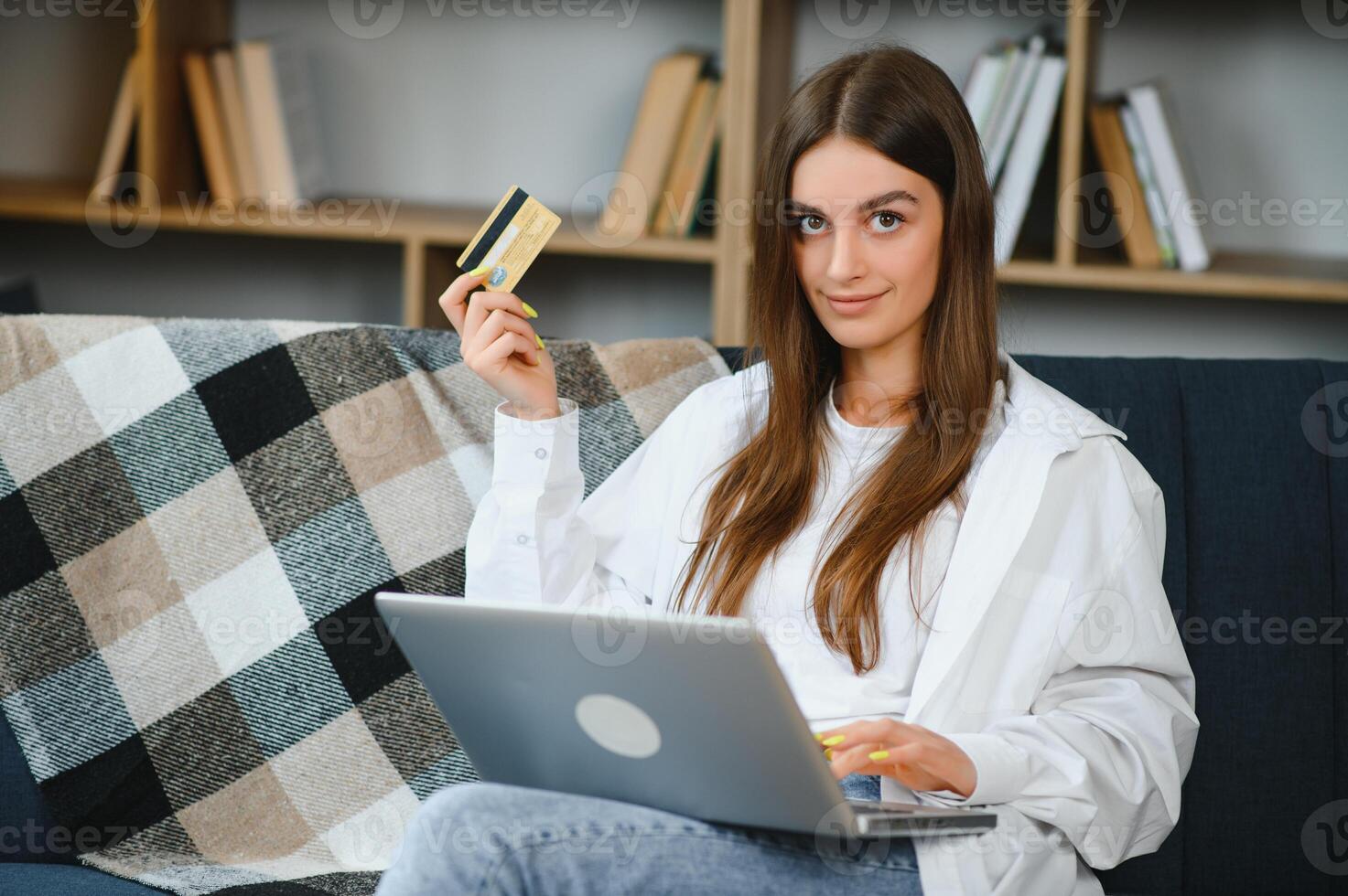 sonriente mujer participación crédito tarjeta y utilizando ordenador portátil en sofá en vivo habitación. foto