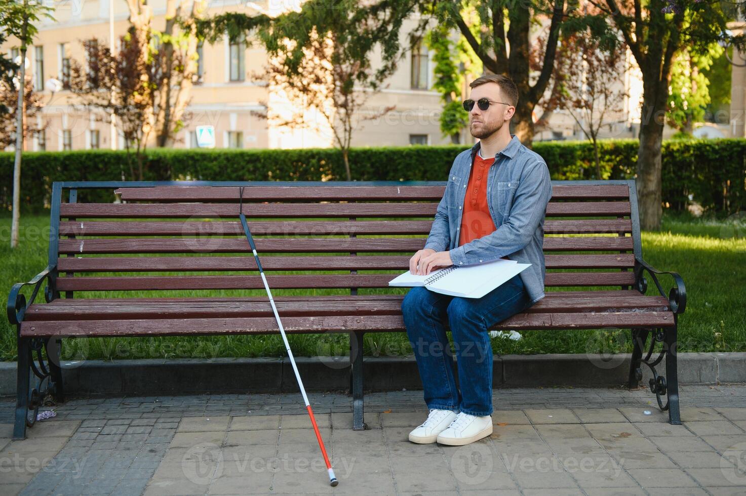 Visually impaired man with walking stick, sitting on bench in city park. Copy space photo