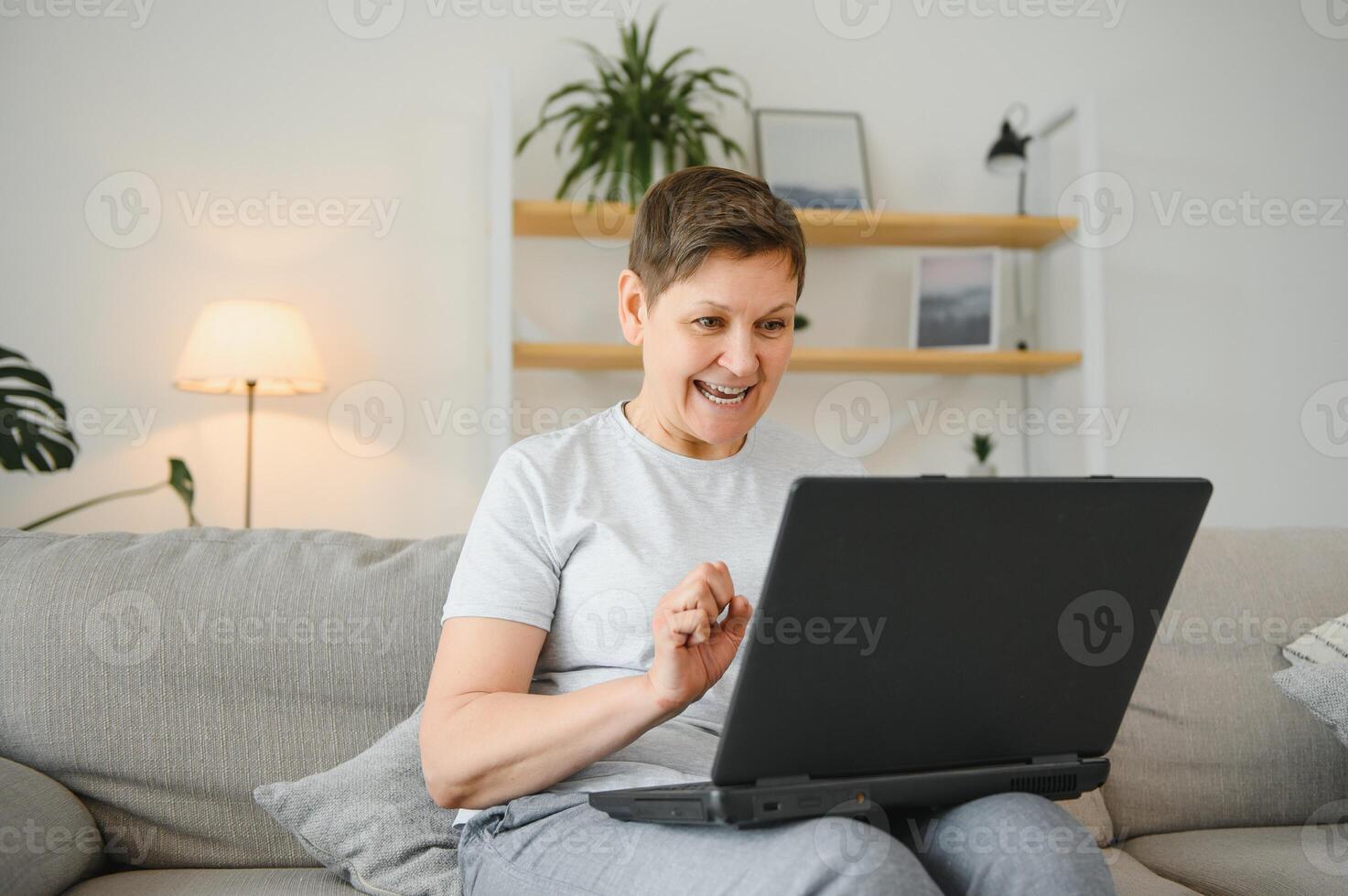 Excited mature woman looking at laptop screen, reading good news in message, celebrating online lottery win, rejoicing success, overjoyed older female sitting on couch at home, using computer. photo