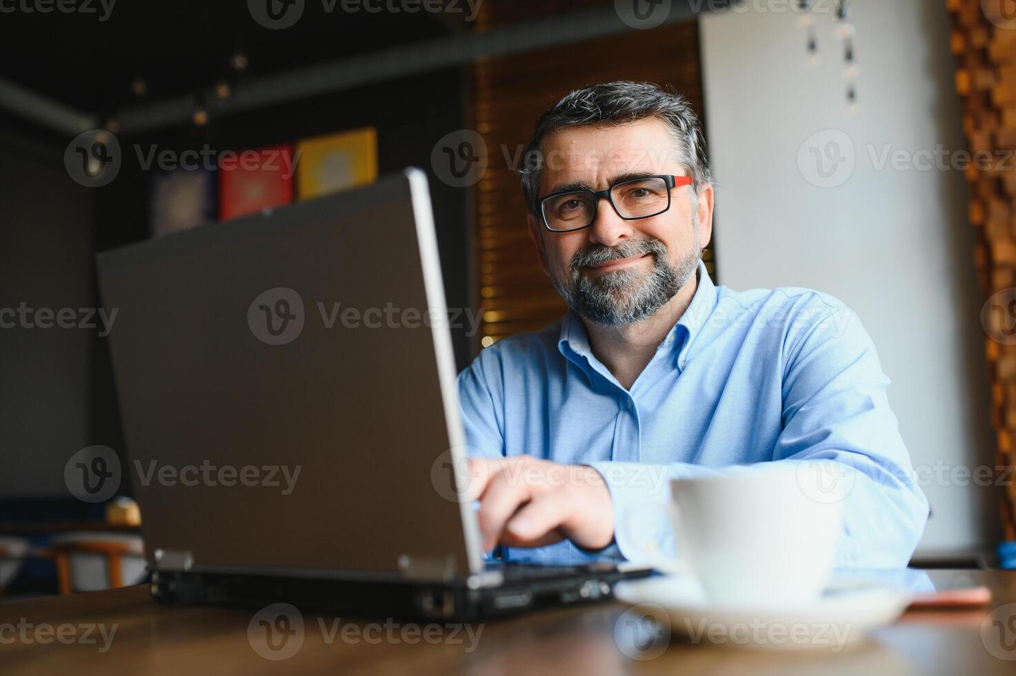 Mature businessman drinking coffee in cafe. Portrait of handsome man wearing stylish eyeglasses using laptop, looking at camera, smiling. Coffee break concept photo