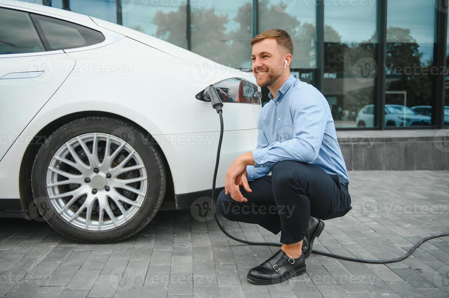 Hansome bearded guy sitting near his new modern electric car and holding plug of the charger, while car is charging at the charging station. photo