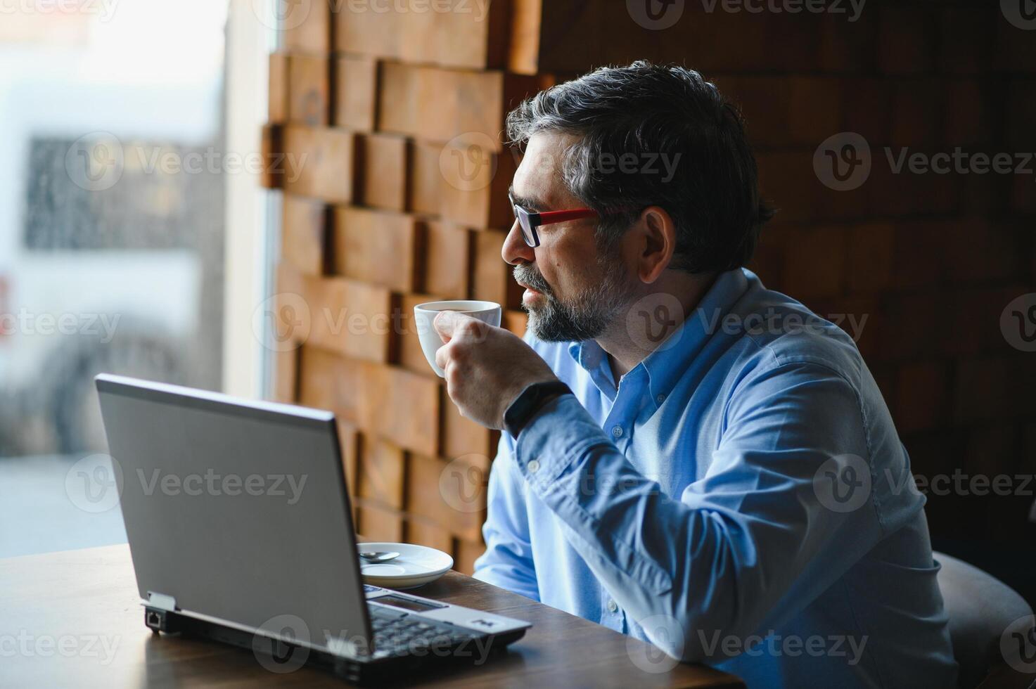 Positive senior bearded man with grey hair drinking coffee and using laptop at cafe, copy space. Stylish aged businessman in burgundy jacket enjoying his tea while working online. photo