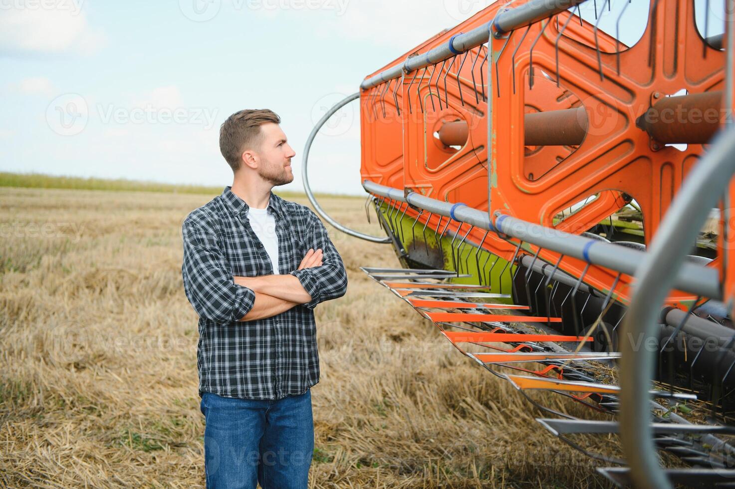 Happy farmer proudly standing in a field. Combine harvester driver going to crop rich wheat harvest. Agronomist wearing flannel shirt, looking at camera on a farmland photo