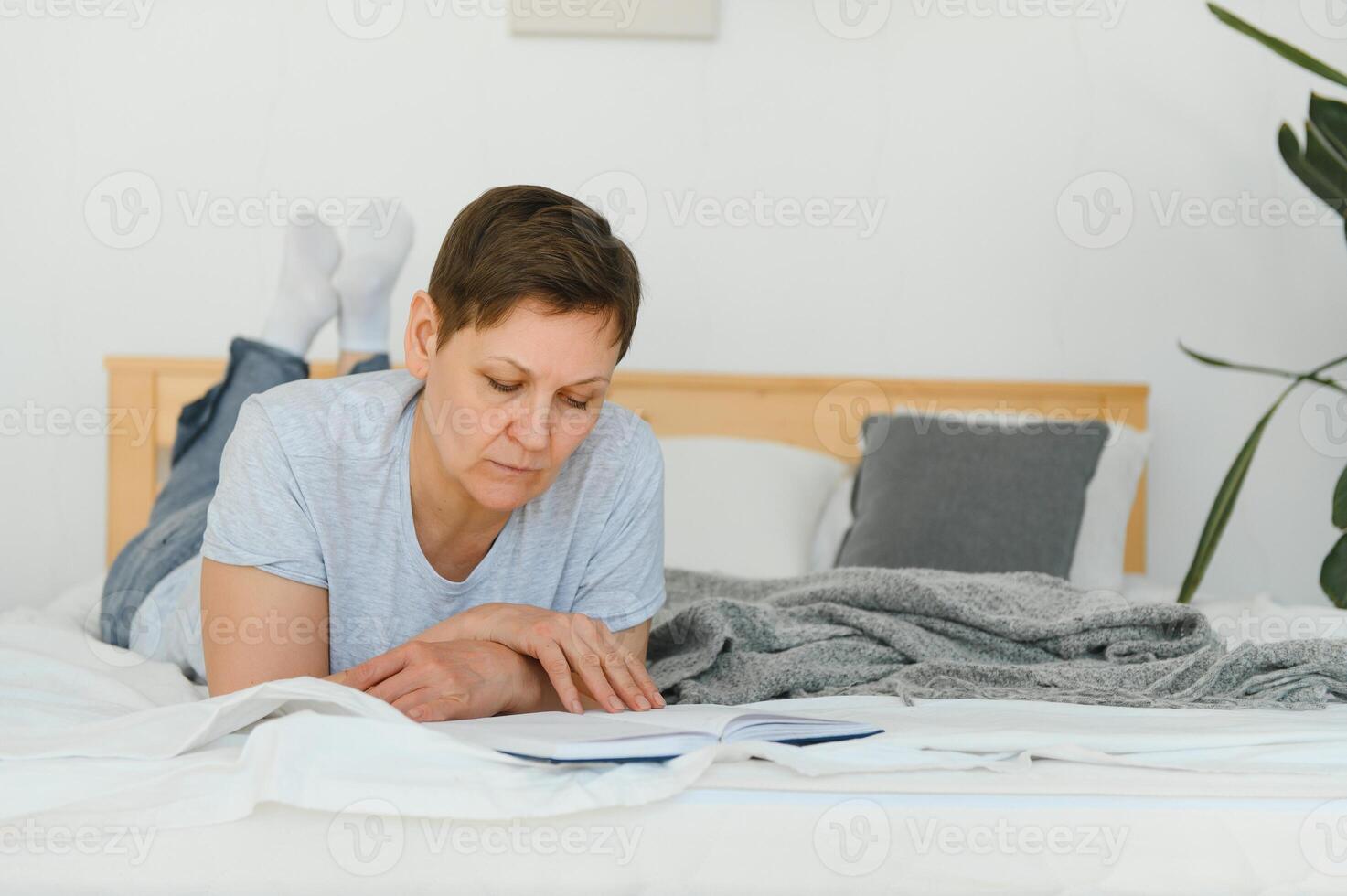 medio años rubia mujer leyendo libro acostado en el cama a hogar. foto