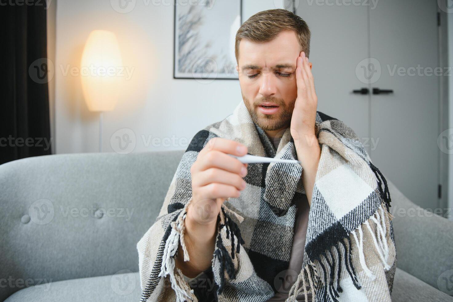 Sick bearded man who has bad cold or seasonal flu sitting on couch at home. Guy with fever wearing warm plaid shivering with worried face expression. photo