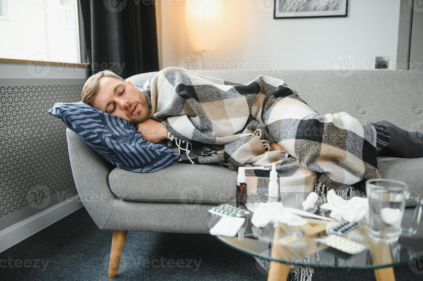 Sick bearded man who has bad cold or seasonal flu sitting on couch at home. Guy with fever wearing warm plaid shivering with worried face expression. photo