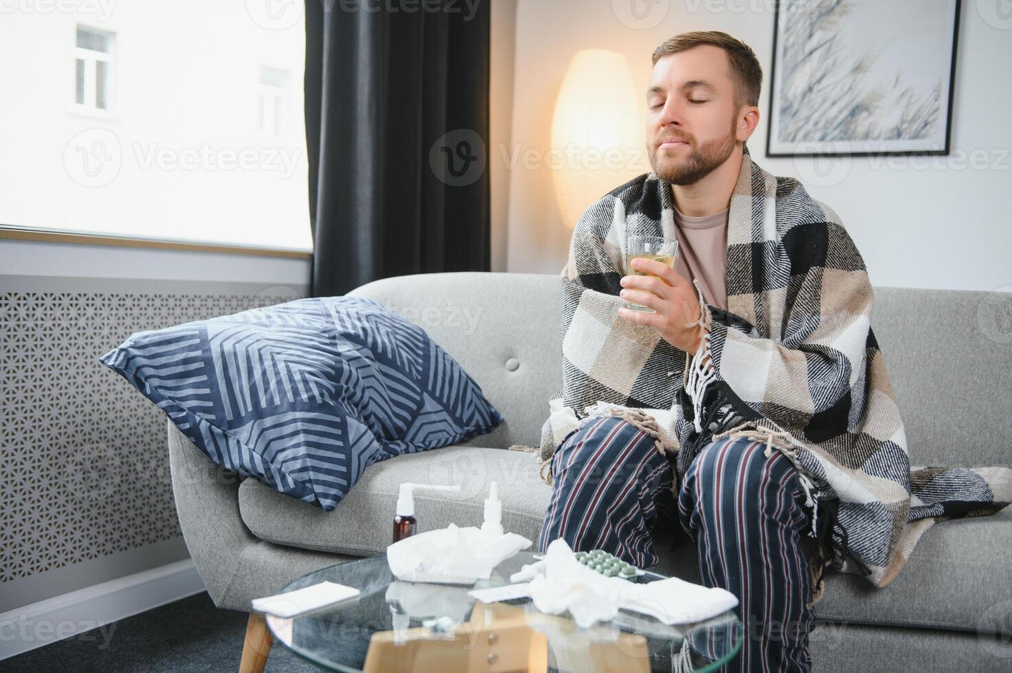 Sick bearded man who has bad cold or seasonal flu sitting on couch at home. Guy with fever wearing warm plaid shivering with worried face expression. photo