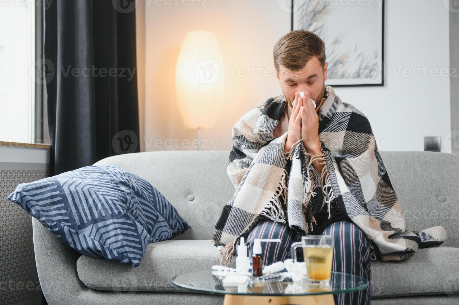 It's cold at home in wintertime. Man freezing in his house in winter because of broken thermostat. young guy wrapped in woolen plaid shivering while sitting on sofa in living room interior photo