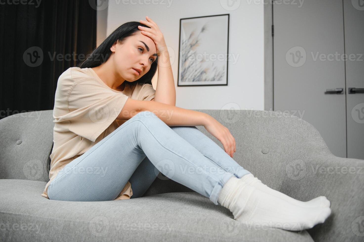 Portrait of beautiful young woman with depressed facial expression sitting on grey textile couch. Cyber bullying victim concept. Sad female in her room. Background, copy space photo