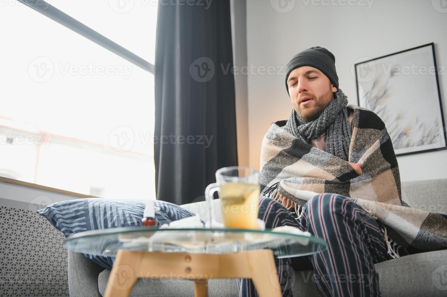 It's cold at home in wintertime. Man freezing in his house in winter because of broken thermostat. young guy wrapped in woolen plaid shivering while sitting on sofa in living room interior photo