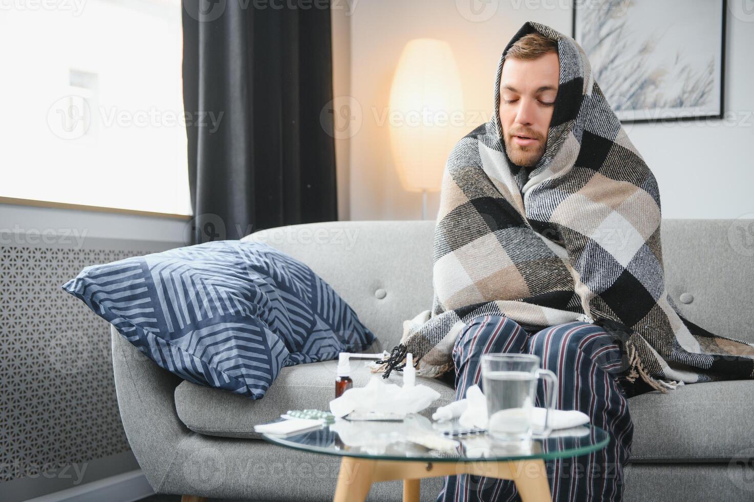 Sick bearded man who has bad cold or seasonal flu sitting on couch at home. Guy with fever wearing warm plaid shivering with worried face expression. photo